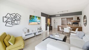 Ein modernes Wohnzimmer mit gelber und weißer Couch sowie einem Essbereich mit weißem Tisch und Stühlen. Im Hintergrund ist eine Küche mit Mikrowelle und Schränken zu sehen.