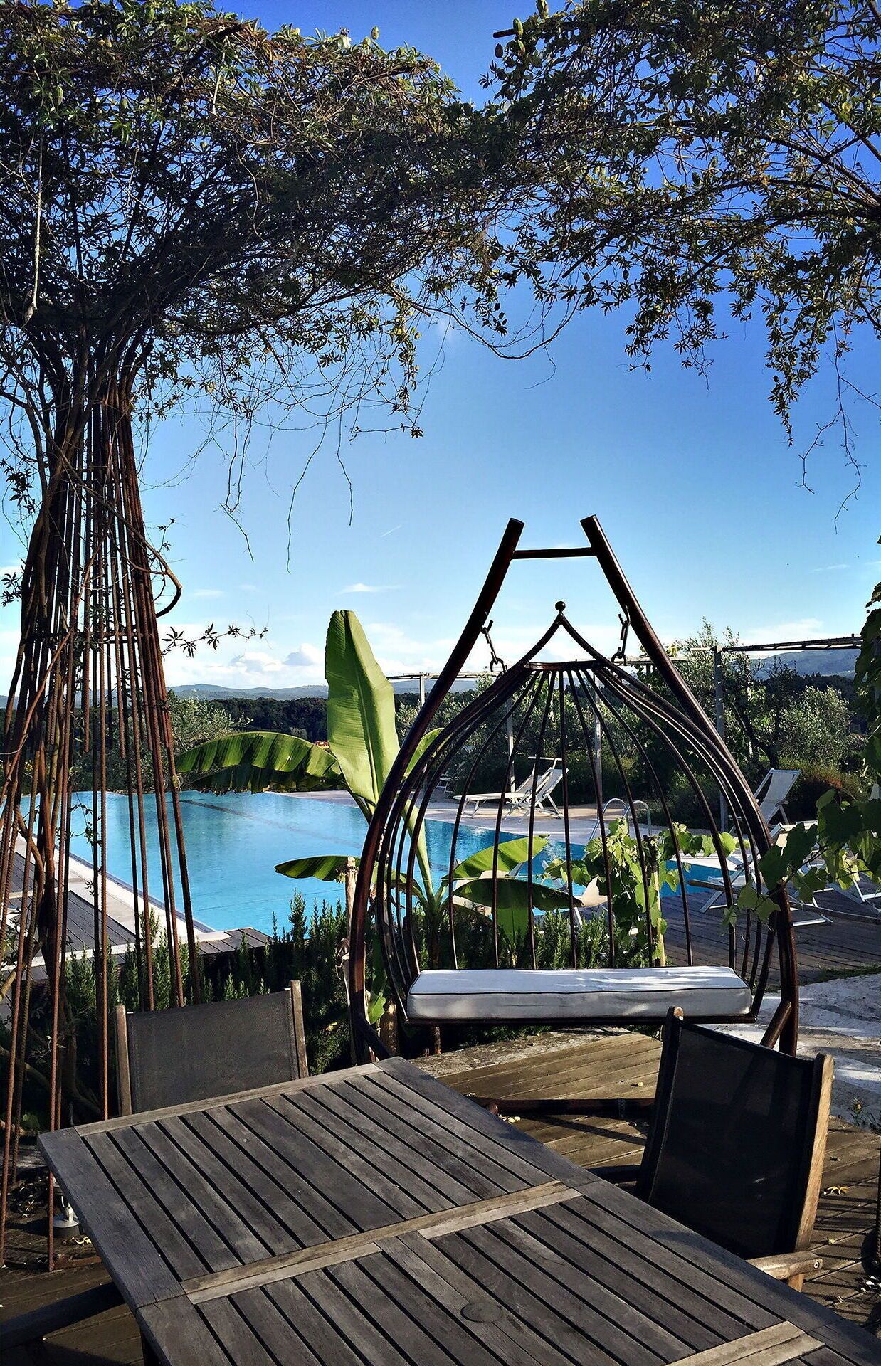 A wooden deck with a swing chair, a hanging chair, and a view of a pool and mountains.