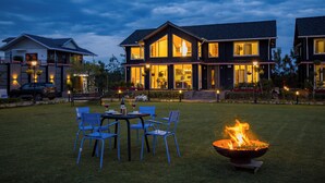 A modern two-story house with a large front porch, a fire pit in the yard, and a dining area with chairs. - ELIVAAS Esh Cottages (Palampur)