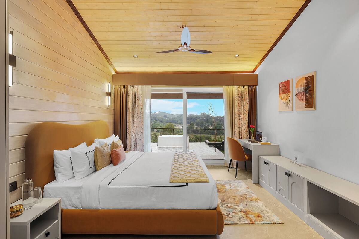 A modern bedroom with a large bed, a desk, and a balcony with a view.