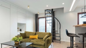 Uma sala de estar moderna com um sofá verde, uma mesa de centro de vidro, uma planta em vaso, uma escada de metal preta e um espaço bar com banquetas.