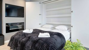 Quarto moderno com cama, televisão embutida e unidade de armazenamento com prateleiras.