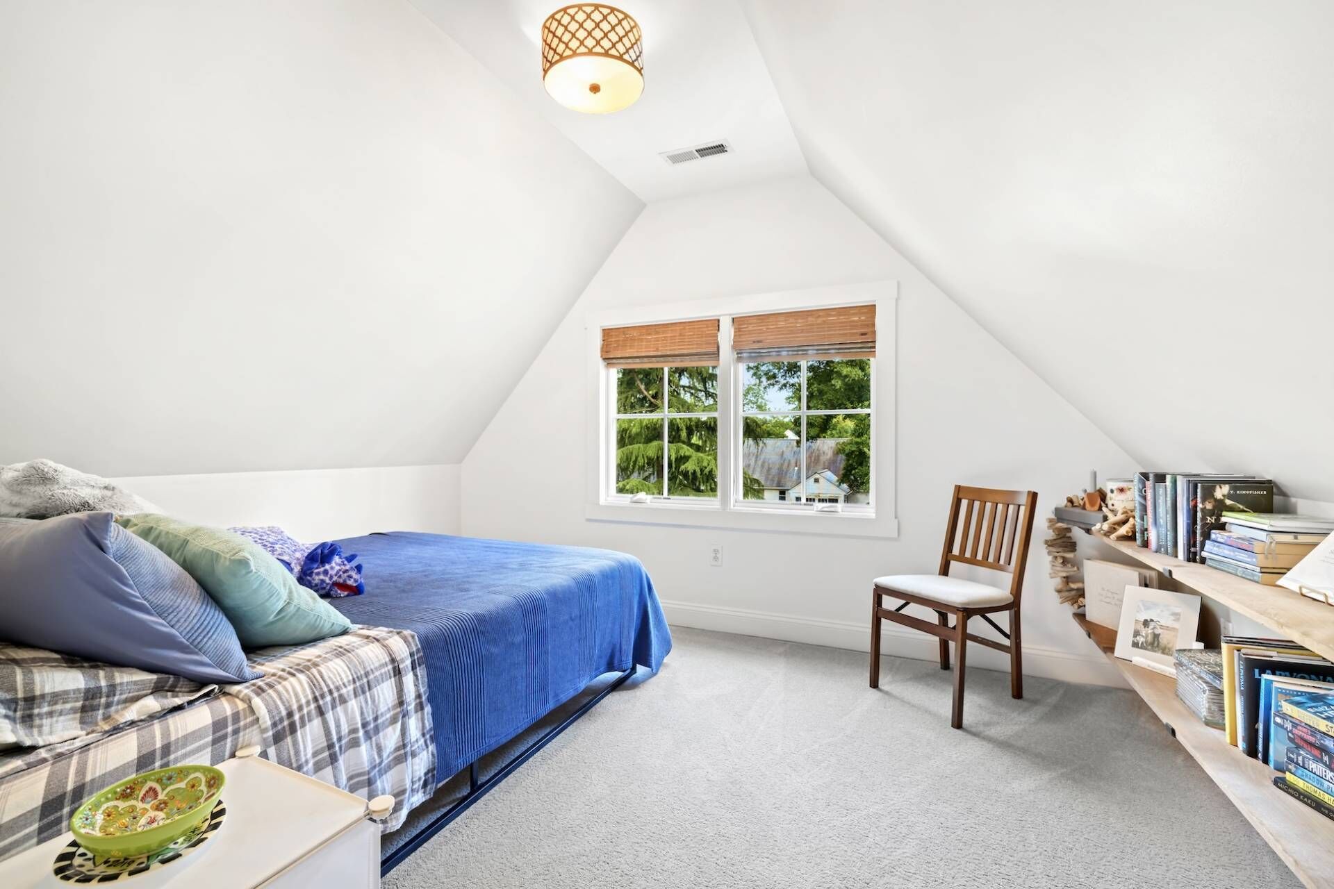 A bedroom with a sloped ceiling, a bed with a blue cover, a chair, a bookshelf, and a window with a view of greenery.