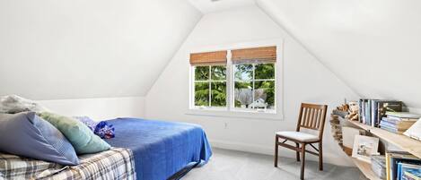 A bedroom with a sloped ceiling, a bed with a blue cover, a chair, a bookshelf, and a window with a view of greenery.