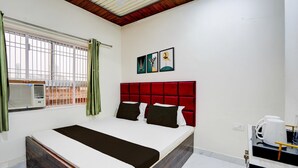 A hotel room with a bed, a window with curtains, a wall-mounted air conditioner, and a coffee maker on a table.