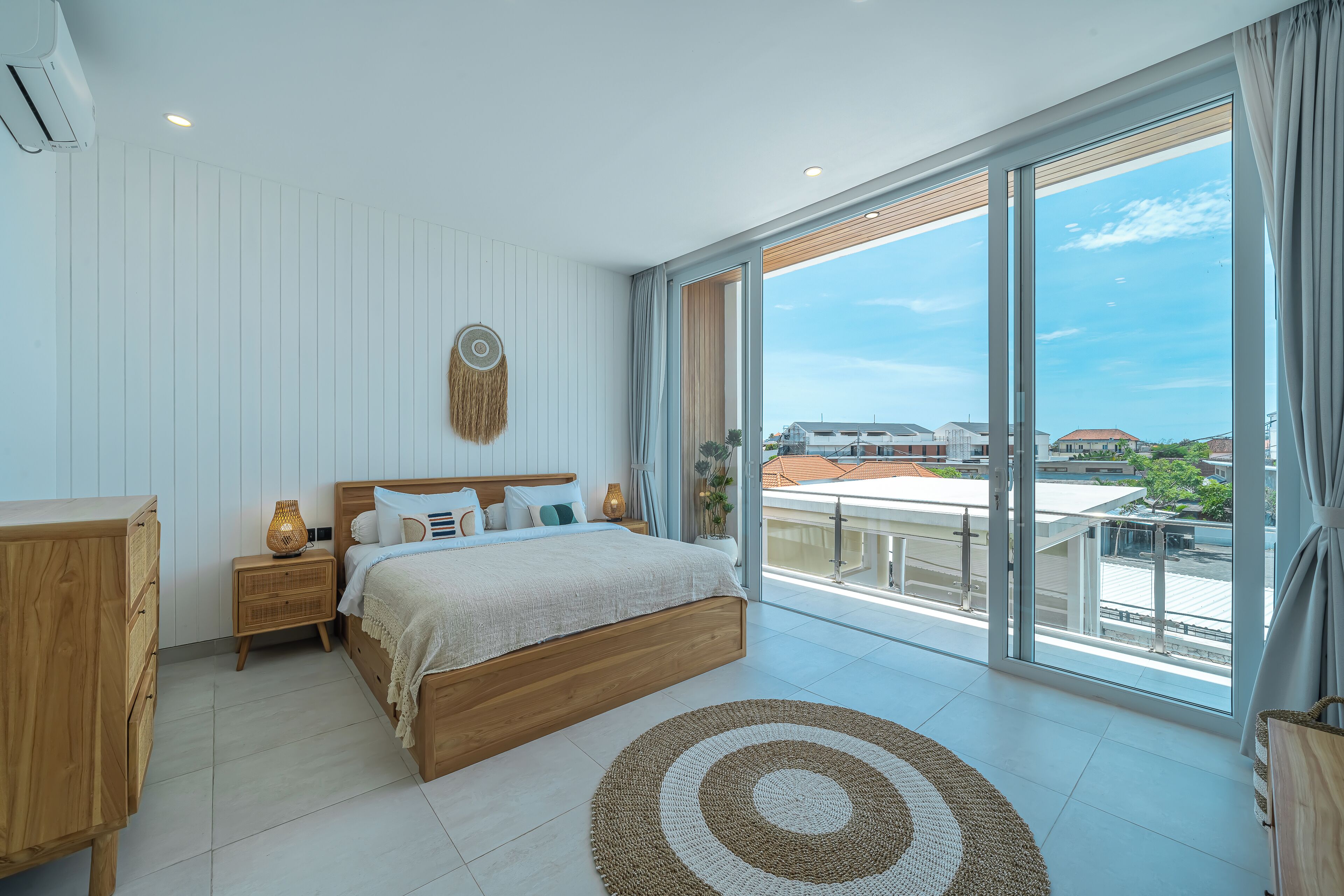 A modern bedroom with a large bed, a nightstand, a wooden dresser, and a view of the city through floor-to-ceiling windows.