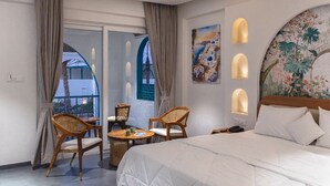 Une chambre d’hôtel avec un lit, deux fauteuils en osier, une petite table et une vue sur un immeuble, visible à travers une porte vitrée.