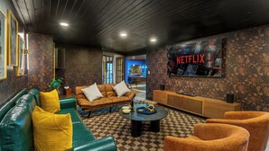 Una sala de estar acogedora con un sofá verde, un sofá de cuero marrón y una mesa de centro. Un cartel grande de Netflix decora la pared, y la habitaci ón tiene un patrón de piso a cuadros.