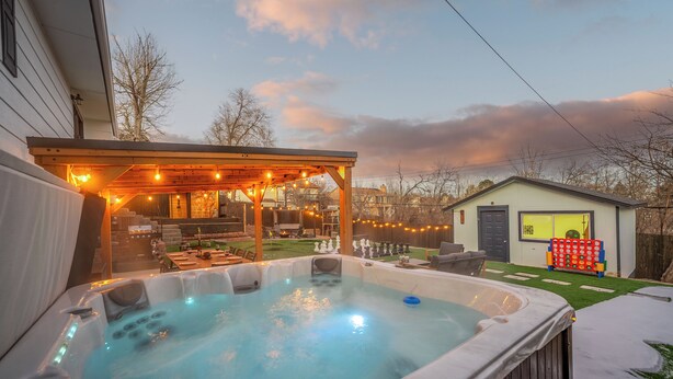 Un jacuzzi con terraza de madera y luces colgantes, rodeado por un patio trasero con un cobertizo pequeño y una zona de juegos.