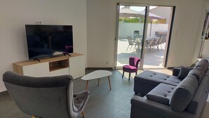 A modern living room with a grey sofa, a grey armchair, a flat-screen TV on a wooden cabinet, a small white table, and a sliding glass door leading to an outdoor patio with chairs and umbrellas. - Villa near Les Sables d'Olonne with private pool 15 minutes from the beaches! (Saint-Mathurin)
