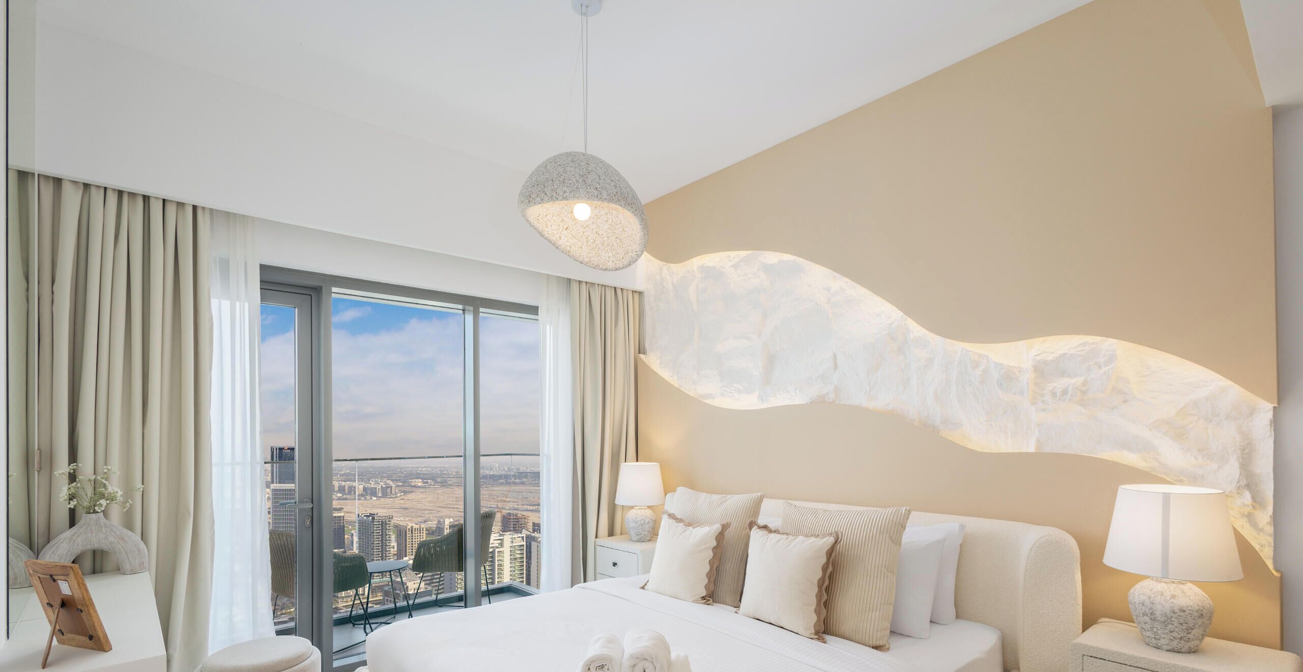 Une chambre d’hôtel moderne dotée d’un grand lit, de deux tables de chevet avec des lampes, d’une vue sur la ville grâce à de grandes fenêtres et d’une œuvre d’art murale originale.