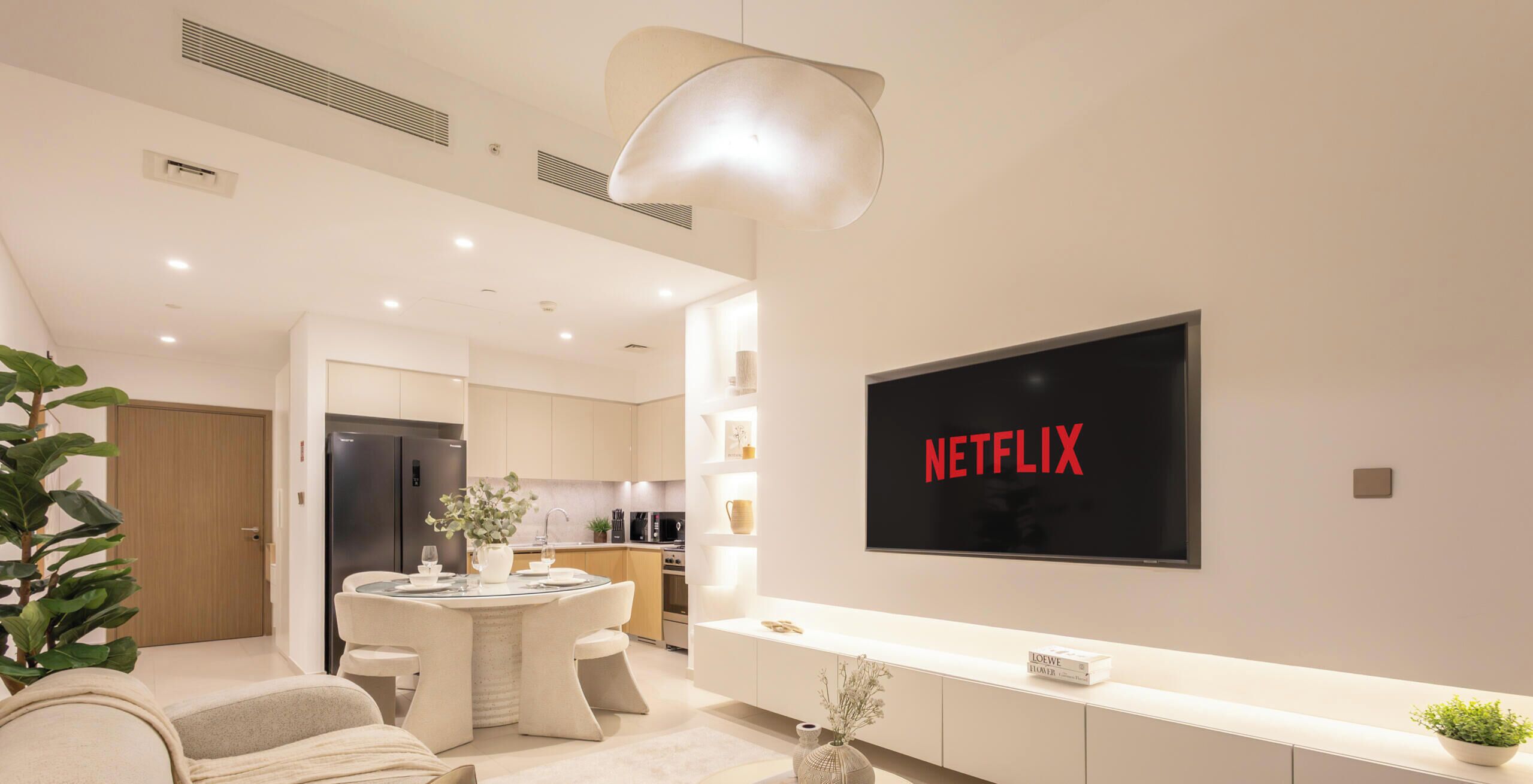 Un salon moderne avec un téléviseur à écran plat fixé au mur, un coin repas avec une table et des chaises, et une cuisine en arrière-plan.