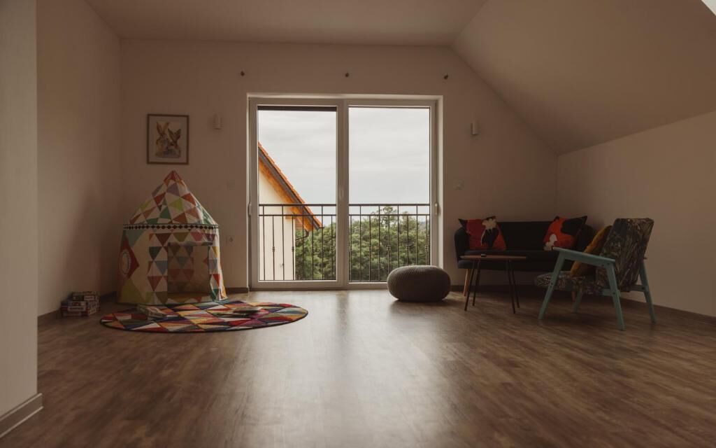 A spacious room with a large sliding glass door leading to a balcony, a colorful play tent, a sofa with cushions, and a patterned rug on the wooden floor.