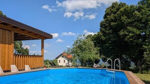 Una piscina con cubierta de madera y sillas de descanso, rodeada de árboles y cielo despejado.