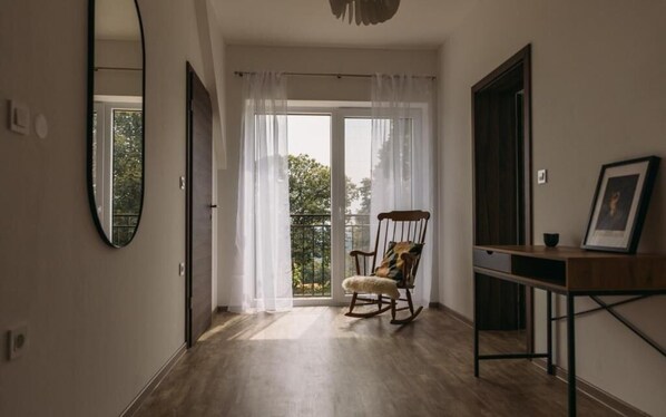 A room with a wooden floor, a rocking chair, a desk with a picture frame, and a large window with sheer curtains. - Hiša na Ravnah (Brežice)