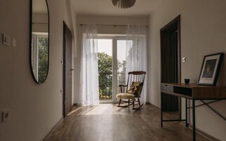 Um quarto com piso de madeira, uma cadeira de balanço, uma escrivaninha com porta-retrato e uma janela grande com cortinas finas.