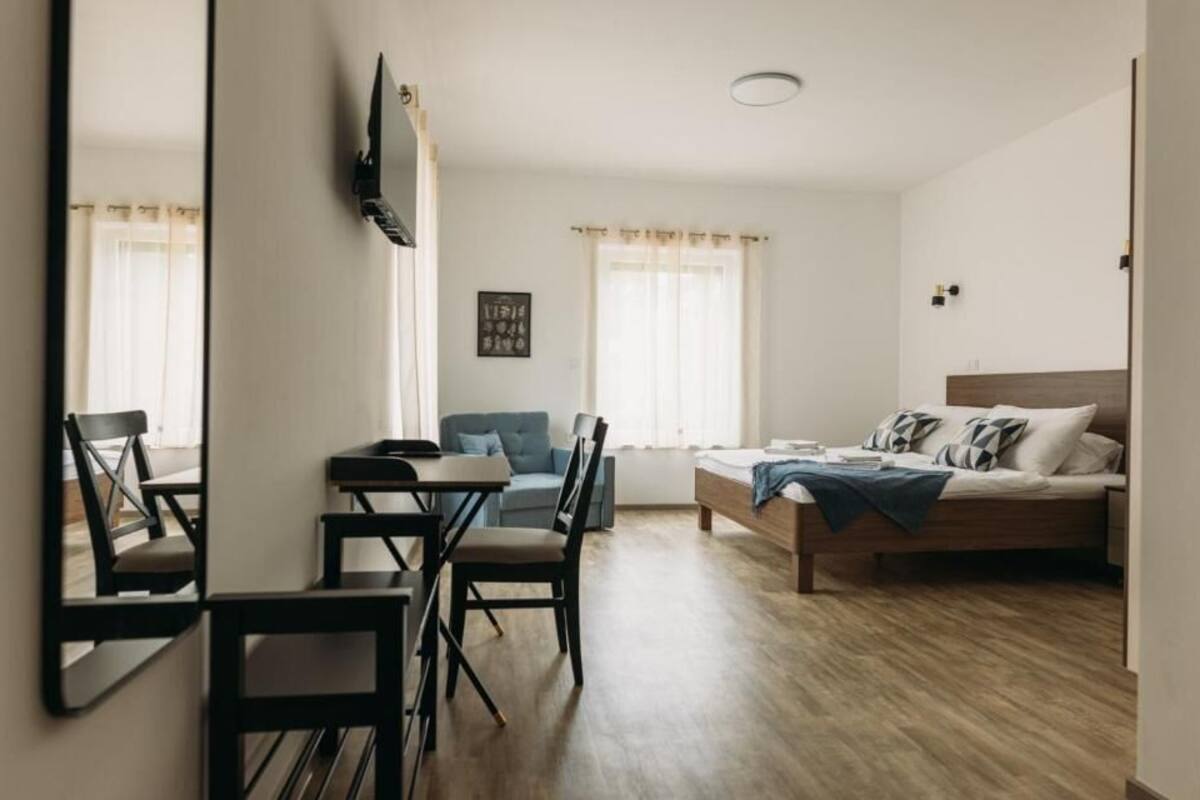 A room with a bed, a desk, and a chair. There is a window with curtains, a wall-mounted TV, and a framed picture on the wall.