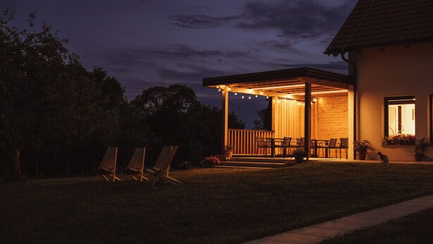 Un espacio de descanso al aire libre cubierto, con luces colgantes, sillas de madera y una planta en maceta.