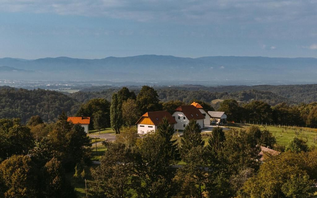 A rural landscape with a large white building with a red roof, surrounded by trees and hills in the background.