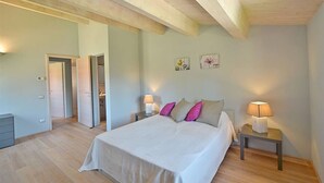 A bedroom with a large bed, two bedside tables with lamps, a door leading to another room, and a light-colored wall with two framed pictures. - Apartment Azzurro- Cignella Resort Tuscany (Trequanda)