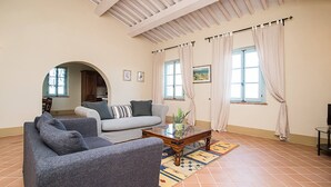 A living room with a sofa, armchair, and coffee table. There are two windows with curtains, a door, and a view into another room. - Apartment Cipresso-Cignella Resort in Tuscany (Siena)