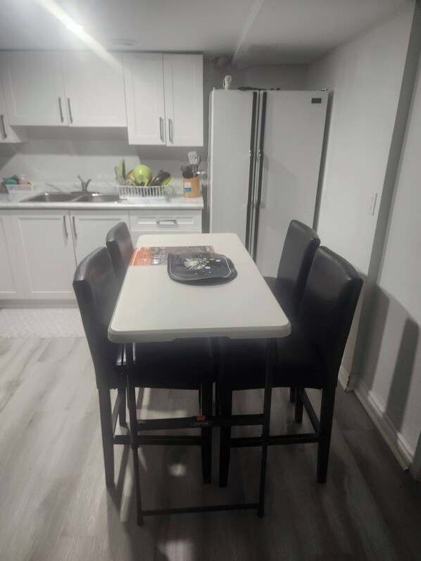 A compact kitchen with white cabinets, a white table, and black chairs. A refrigerator and a sink are visible.