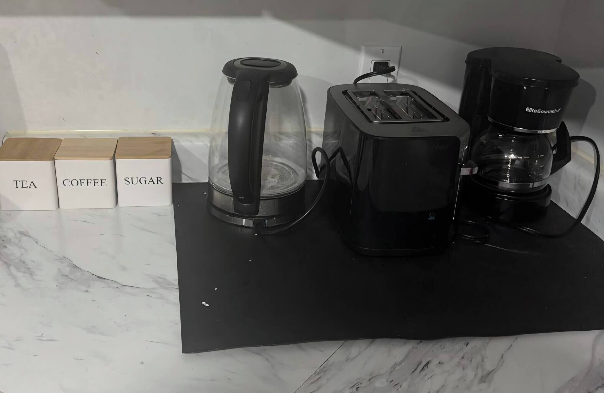 Una encimera de cocina con cafetera, tostadora y licuadora. Hay recipientes etiquetados para té, café y azúcar.
