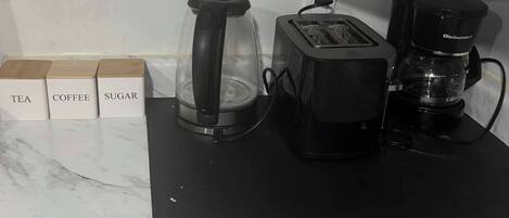 Una encimera de cocina con cafetera, tostadora y licuadora. Hay recipientes etiquetados para té, café y azúcar.