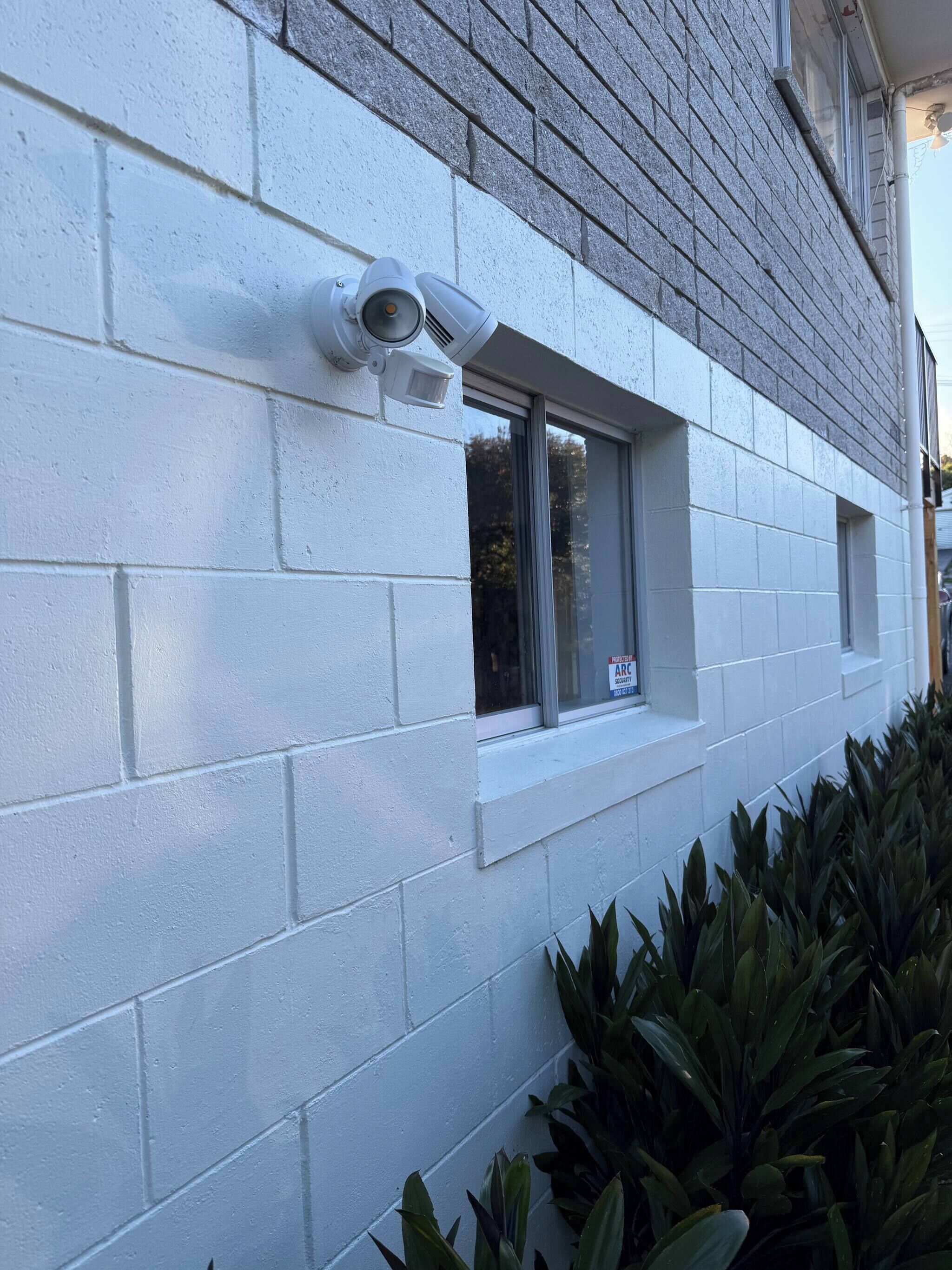 A building with a white brick exterior, a window with a 'For Sale' sign, and a security camera.