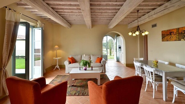 Un vaste salon doté d’un haut plafond en bois, d’un canapé blanc agrémenté de coussins orange, d’une table basse avec une plante, et d’un coin repas avec des chaises et une table blanches.