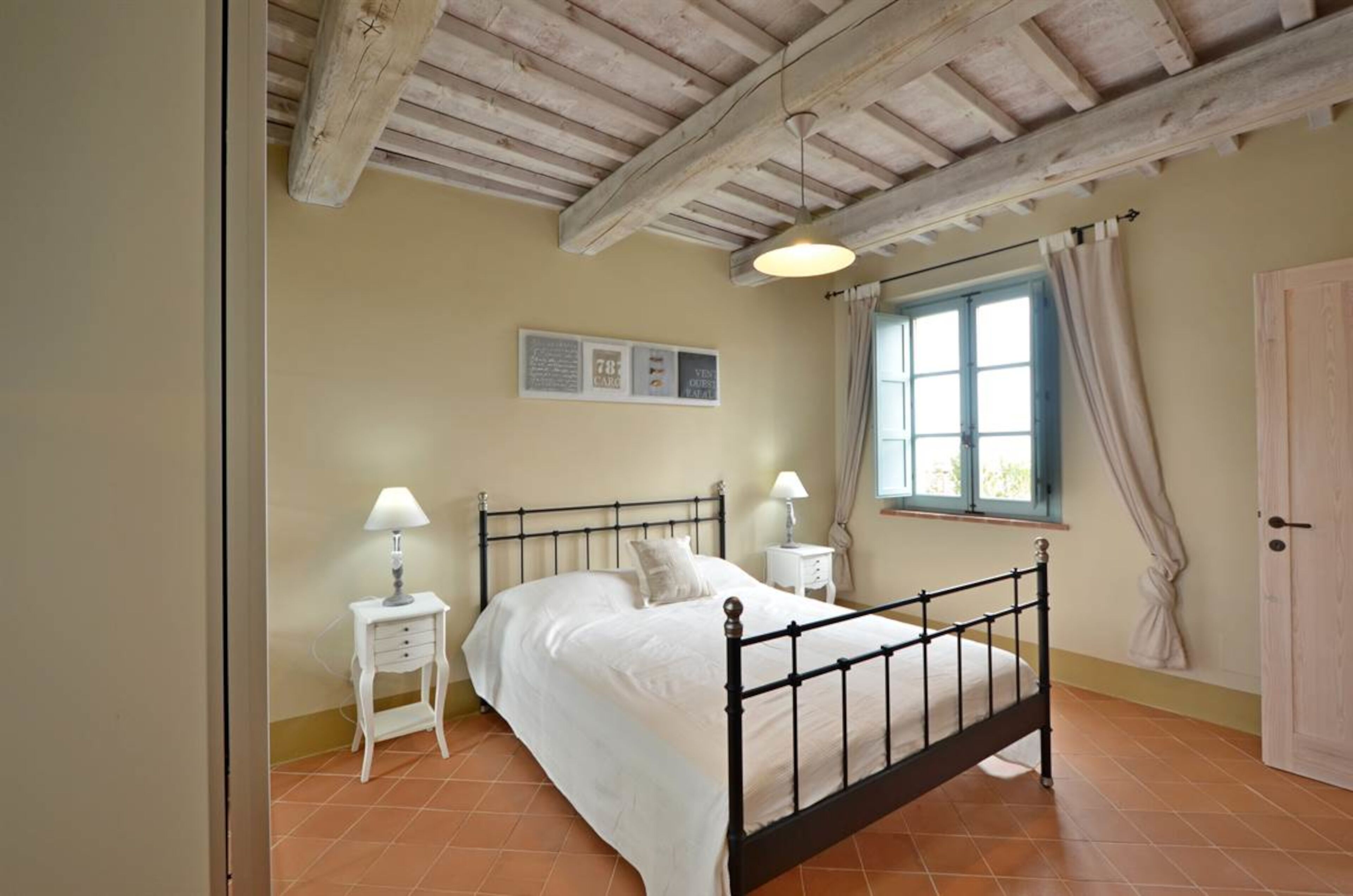 Quarto com cama, mesas de cabeceira, janela com cortinas e teto de madeira.
