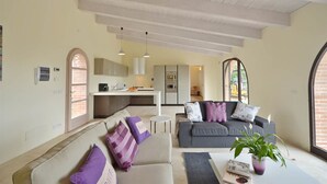 Uma sala de estar moderna com um sofá cinzento, um sofá bege e uma mesa de centro. É possível ver uma cozinha ao fundo, com bancada e armários brancos.