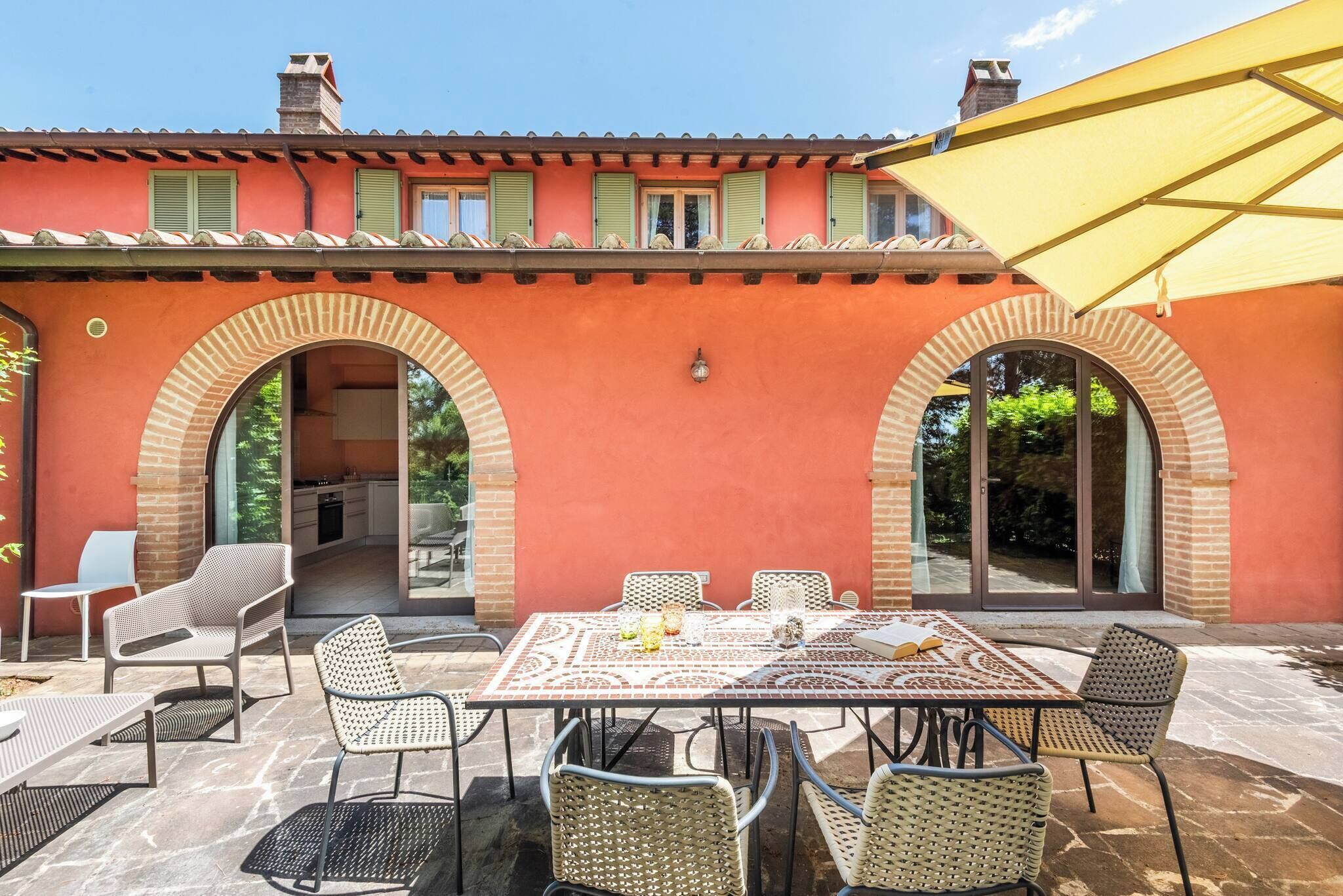 Une terrasse extérieure avec une table et des chaises, un parasol jaune et un mur de briques agrémenté d’arcades.