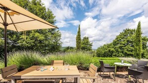 Une terrasse extérieure avec une table et des chaises en bois, un parasol beige et une vue sur un jardin agrémenté d’arbres et de lavande.