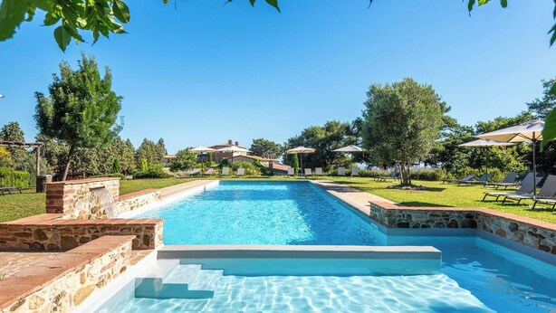 Une piscine rectangulaire avec des marches en pierre menant à l’intérieur, entourée d’une terrasse en pierre et d’une zone herbeuse agrémentée de chaises longues et de parasols.