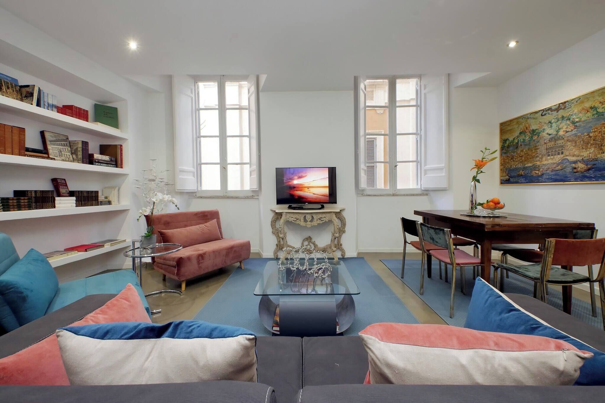 A modern living room with a sofa, coffee table, and TV. There are bookshelves, a dining table, and chairs.