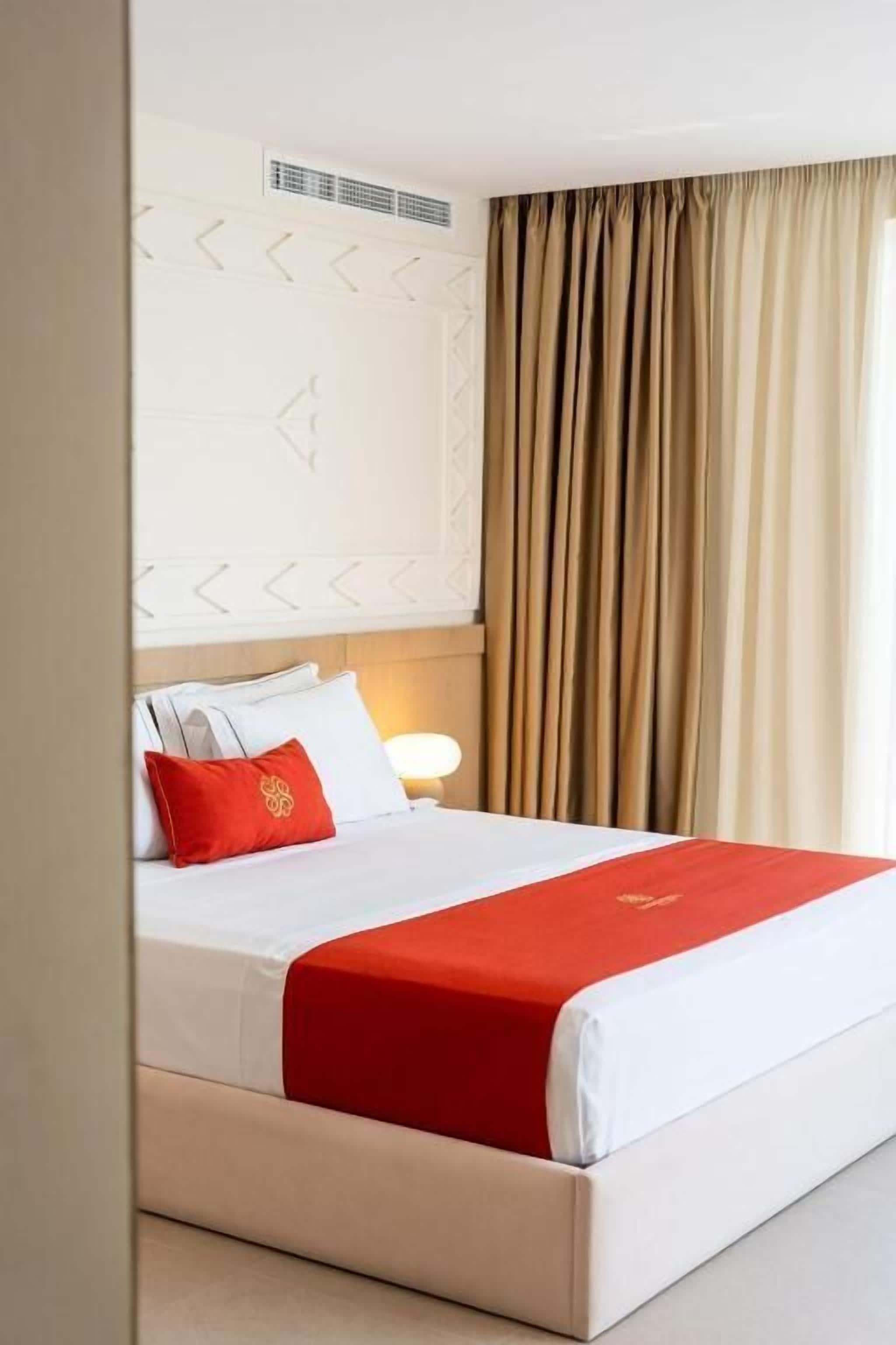 Habitación de hotel con cama, paredes color crema, almohada decorativa roja y ventana con cortinas.