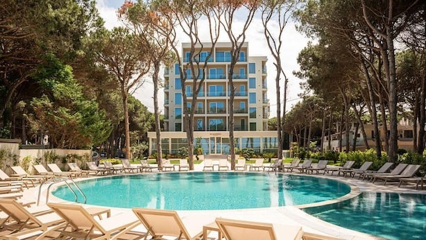 Une piscine entourée de chaises longues, bordée d’arbres, avec un bâtiment visible au loin.