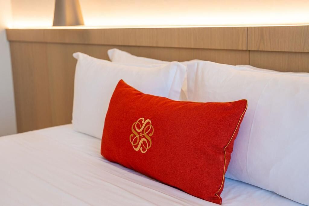 A bed with white bedding and a single red pillow with a gold emblem.