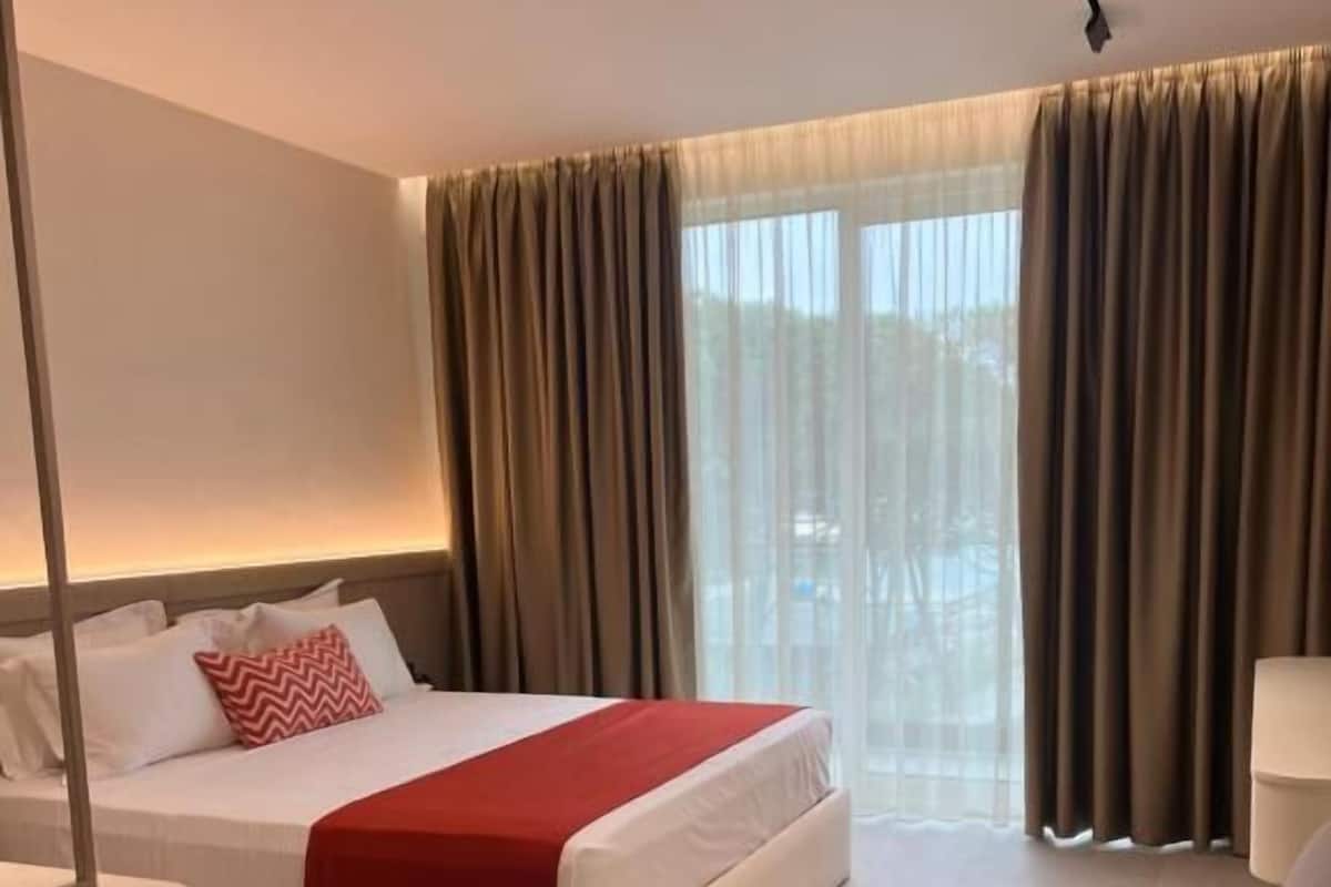 A modern hotel room with a large bed, a sofa, and a view of the outdoors through sheer curtains.