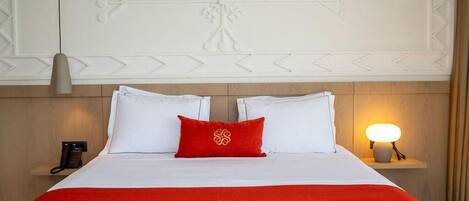 A hotel room with a neatly made bed, a red and white bedspread, a red pillow, a bedside lamp, and a telephone on a nightstand.