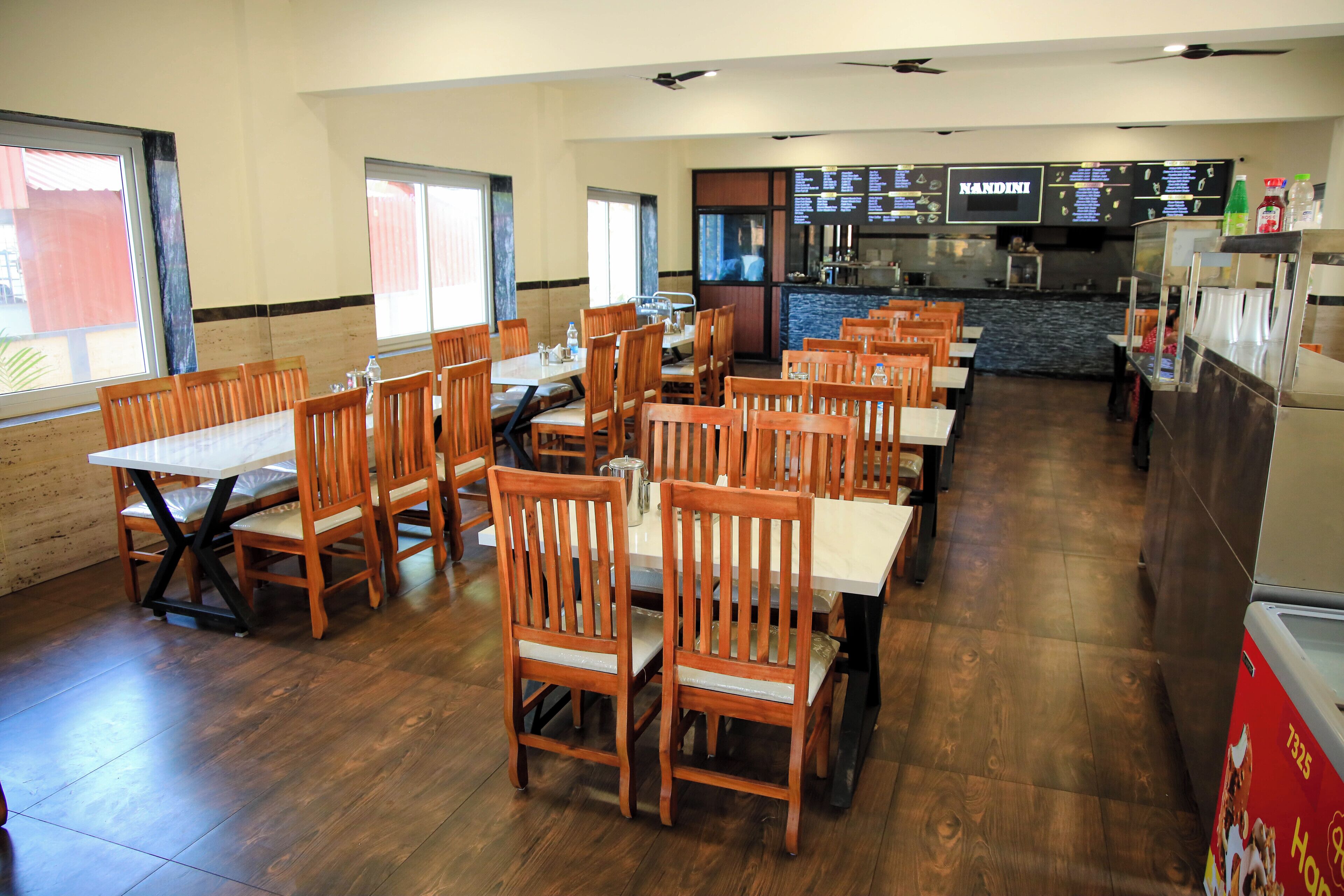 A restaurant with wooden tables and chairs, a kitchen area, and a menu board.