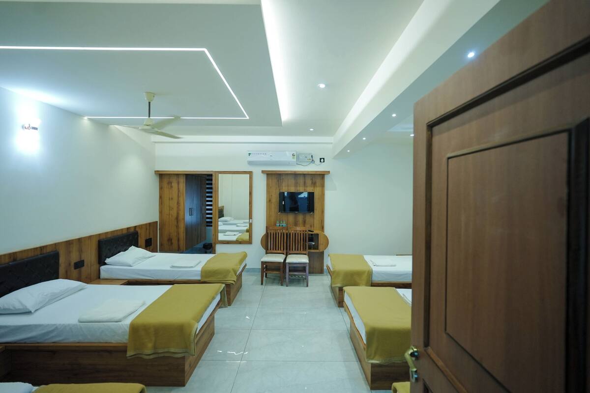 A hotel room with four single beds, a desk with a chair, a TV mounted on the wall, and a ceiling fan.