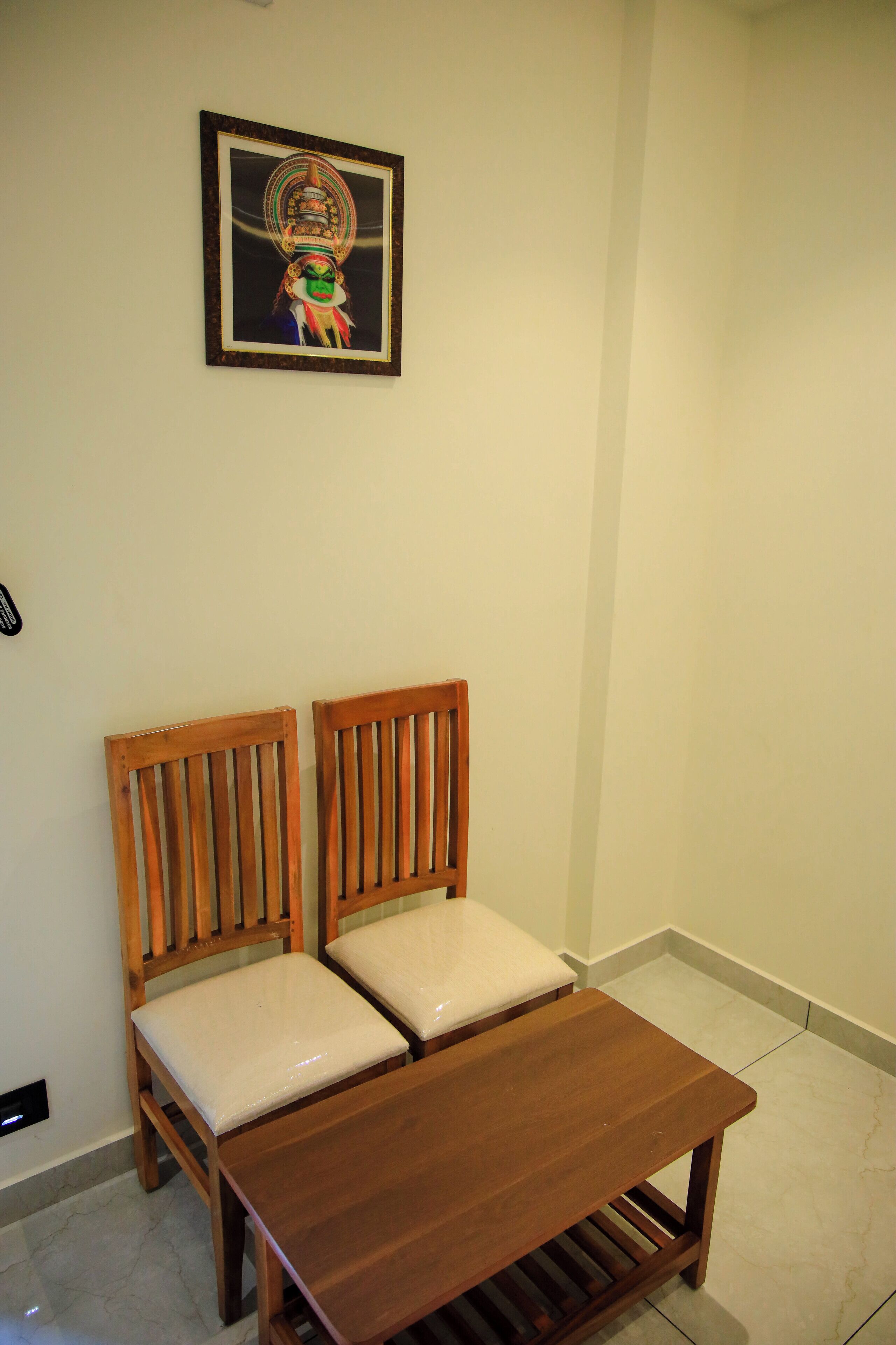 Een kamer met twee houten stoelen met beklede zitplaatsen, een klein houten tafeltje en een lijst met een afbeelding aan de muur.