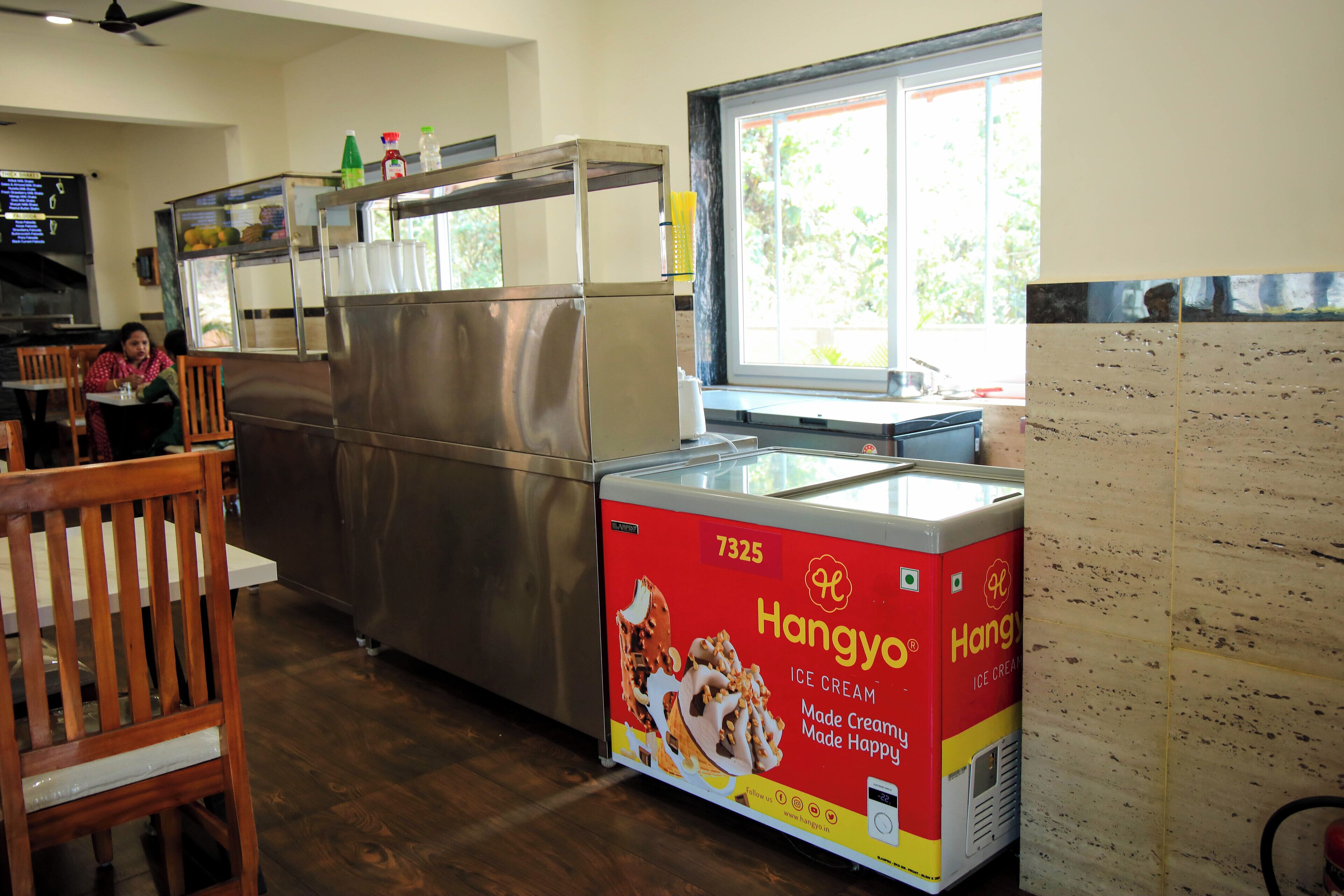 A restaurant interior with a large ice cream freezer, wooden chairs, and a window with a view of greenery.