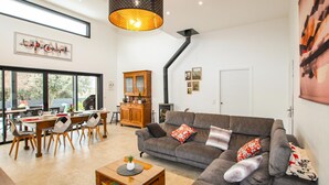 Ein großzügiger Wohnbereich mit einer großen, grauen Couch, einem Holztisch und einem Esstisch mit Stühlen. Es gibt eine Küchenzeile mit Herd und Schränken, und ein großes Fenster sorgt für natürliches Licht.