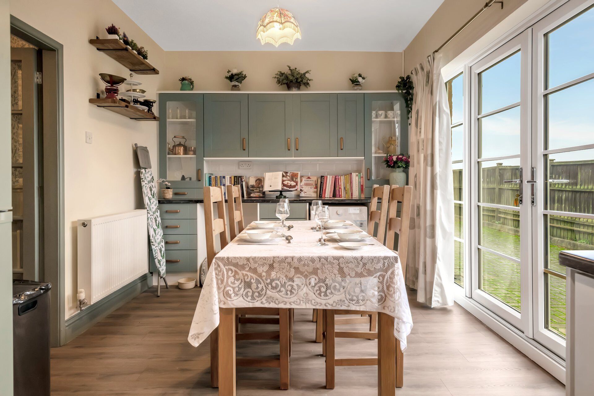 Un coin repas avec une table dressée pour quatre personnes, une cuisine en arrière-plan et une vue sur un jardin par de grandes fenêtres.
