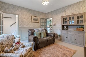 A living room with a brown leather sofa, patterned armchair, and a rug. There is a cabinet with glass doors and a teddy bear on the armchair. - Pools Cottage 2-Head Cooks Cottage in Warwickshire (Kenilworth)