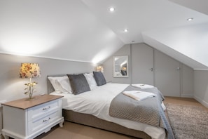 A bedroom with a sloped ceiling, a bed with a grey quilt, a bedside table with a lamp, and a mirror on the wall. - Pools Cottage 2-Head Cooks Cottage in Warwickshire (Kenilworth)