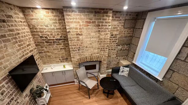 Ein kompakter Wohnraum mit Backsteinwand, einem Flachbildfernseher, einer Couch, einem Sessel, einem kleinen Tisch und einem Fenster mit Aussicht.